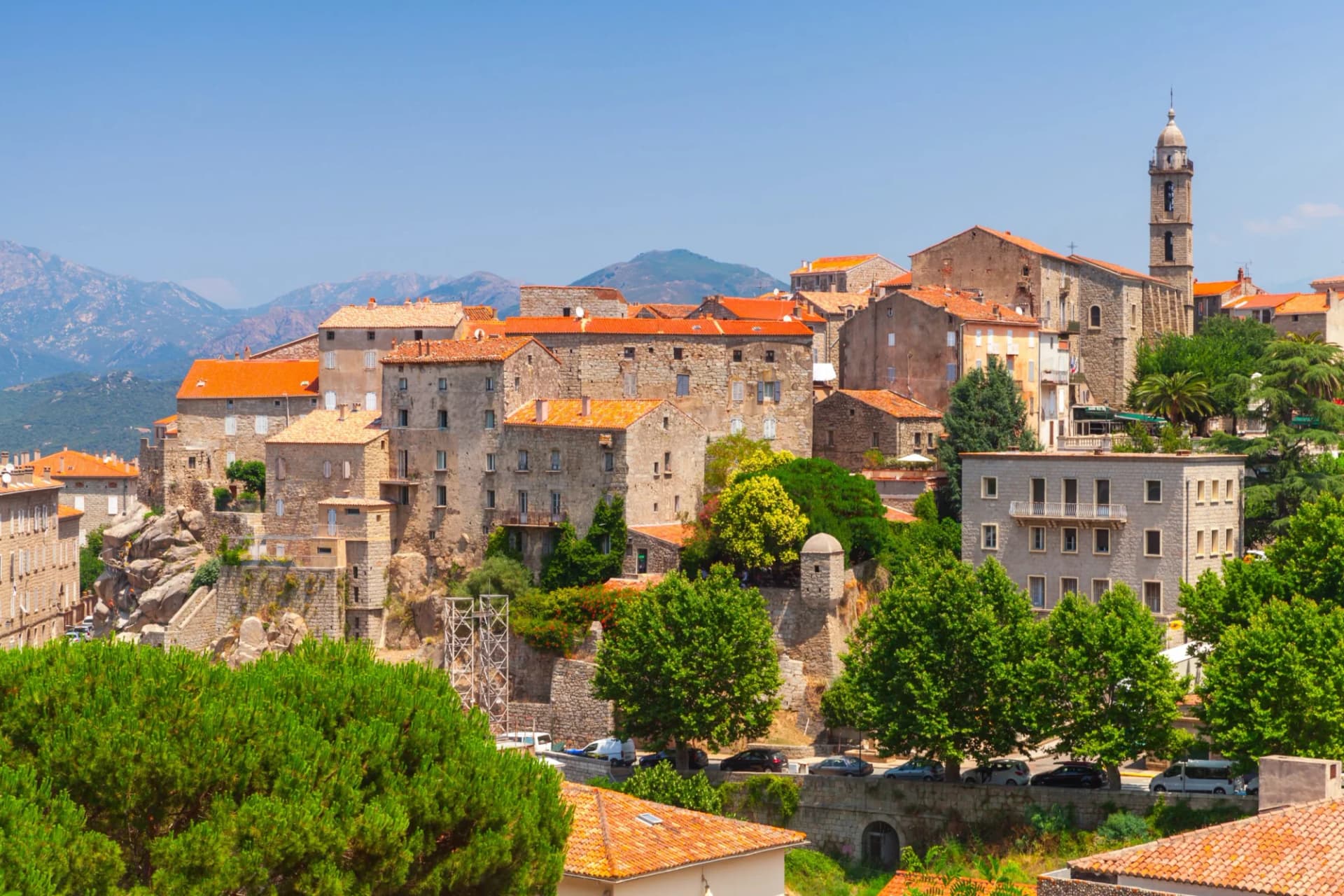 Old town buildings with terracotta roofs climbing a hillside below mountains in Sartene, Corsica.