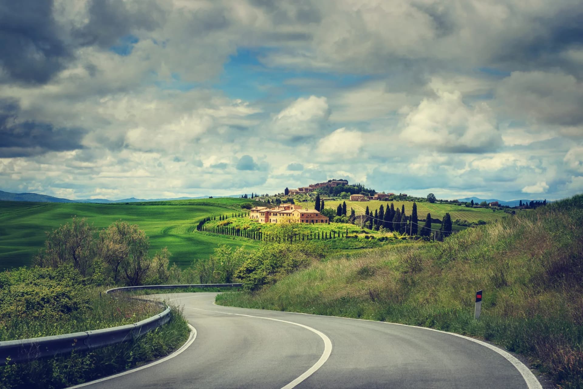 Winding road through green rolling hills toward a Tuscan villa under cloudy skies