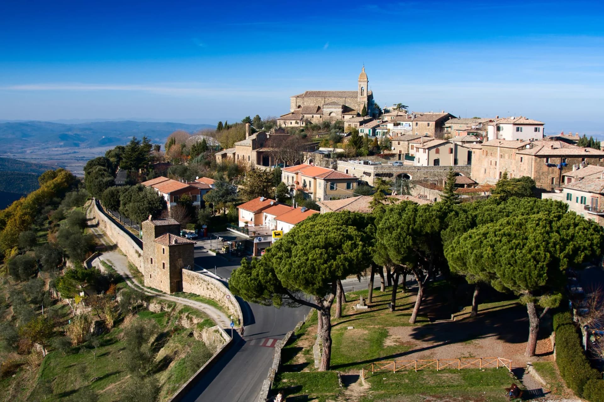 Hilltop town with stone buildings, church tower, and pine trees overlooking valley, Montalcino.