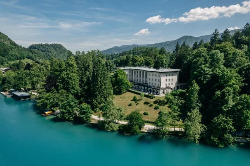 Large white building on wooded hillside overlooking bright turquoise alpine lake, Vila Bled, Slovenia
