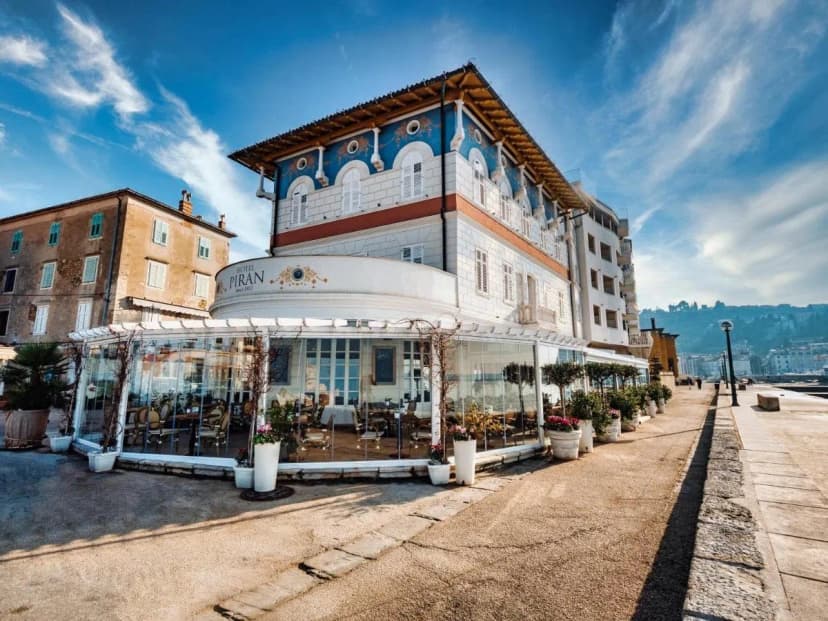 Hotel Piran building with glass-enclosed dining area next to a seaside promenade.