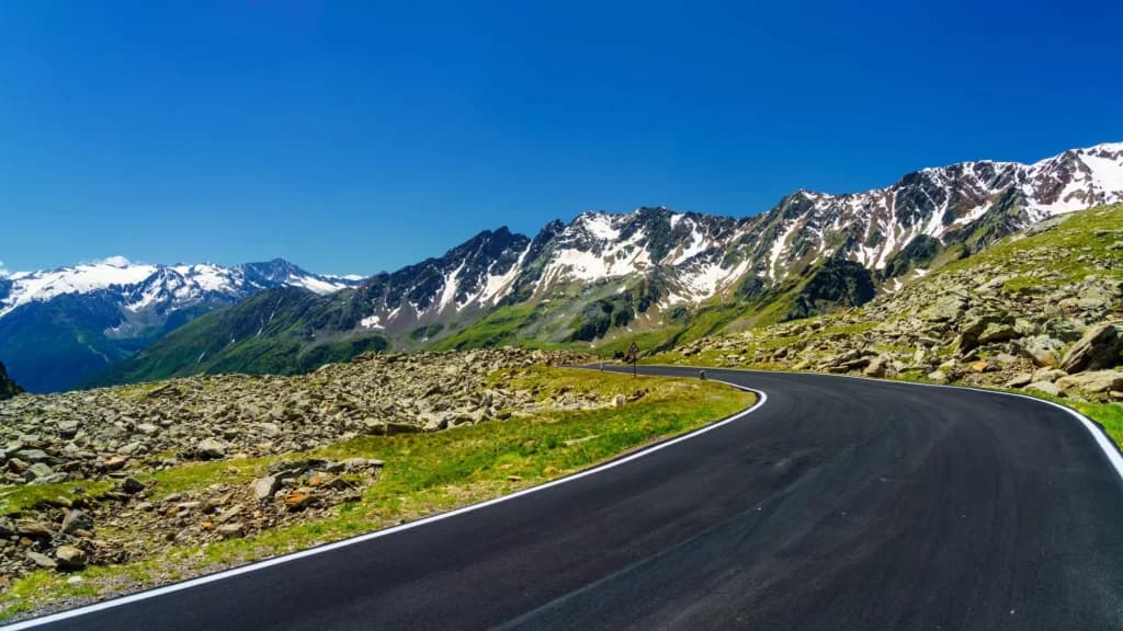 Winding mountain road next to rocky terrain with snow-capped peaks under a clear blue sky.