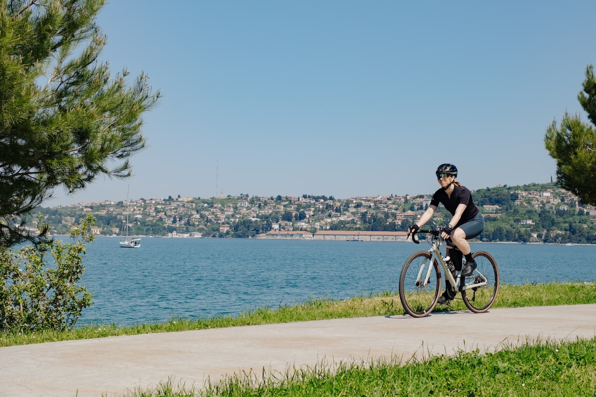 Cyclist riding along a paved path by the water with a town on the distant hillside.