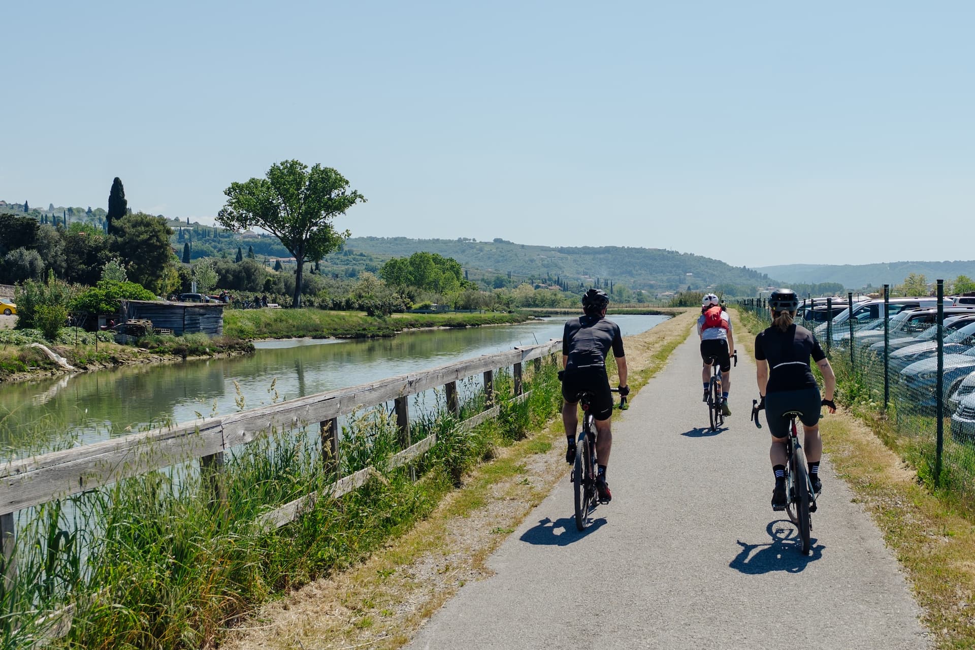 Cyclists riding on a paved path alongside a canal with rolling green hills in the background.