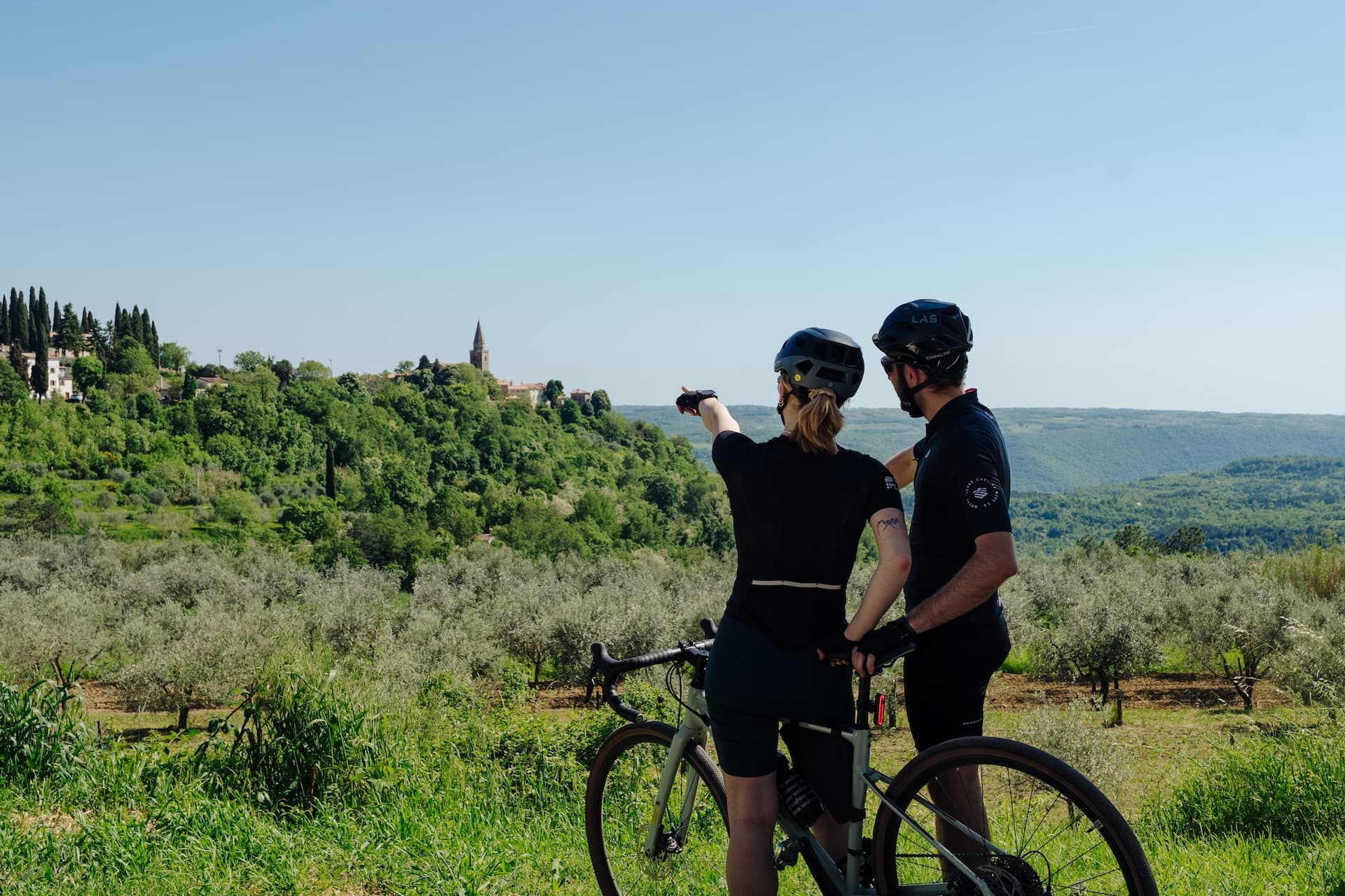 Cyclists pause with a bicycle overlooking an olive grove toward a hilltop village in Grožnjan.