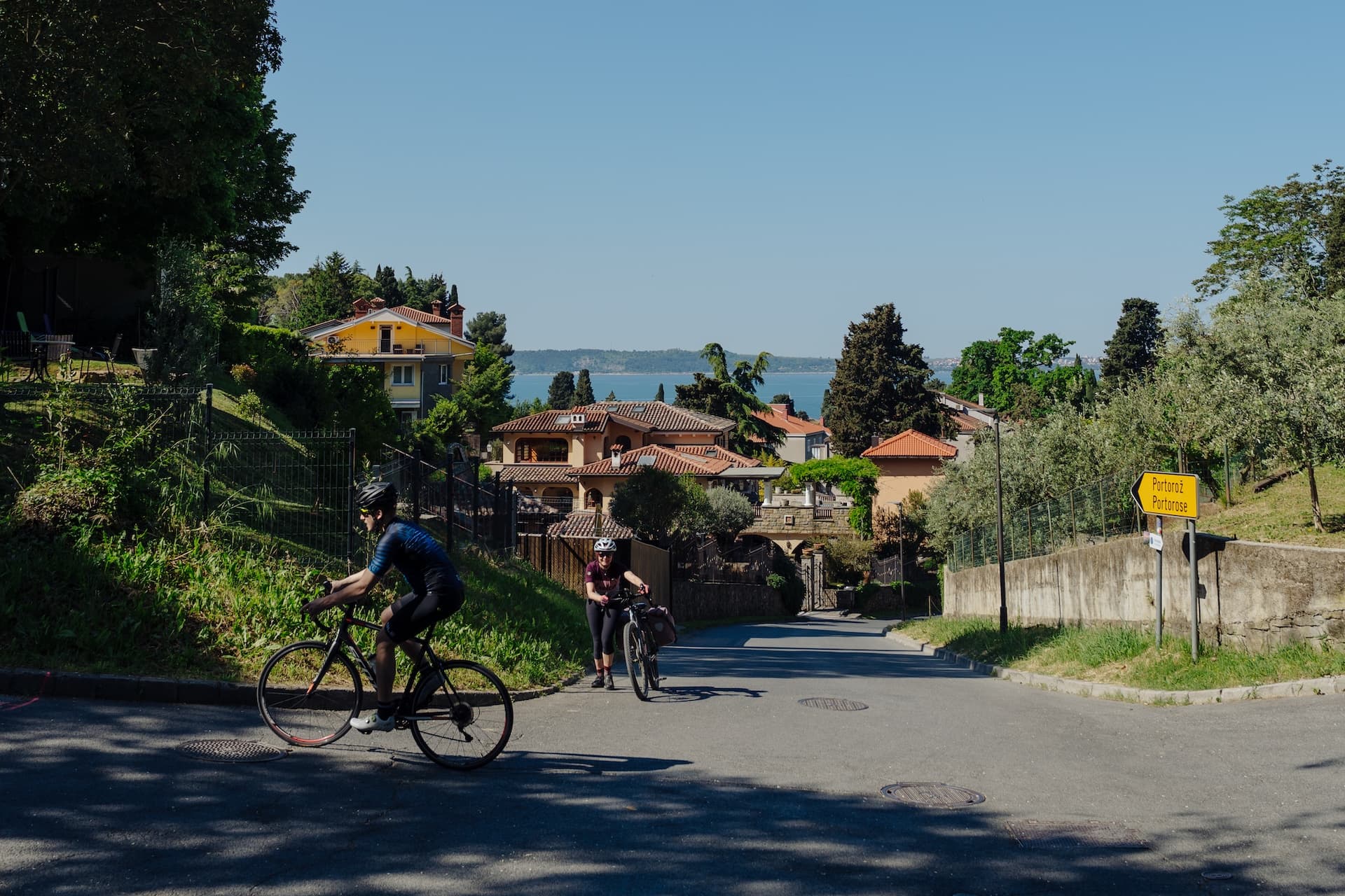 Cyclists on paved road near coastal villas with Portorož sign visible