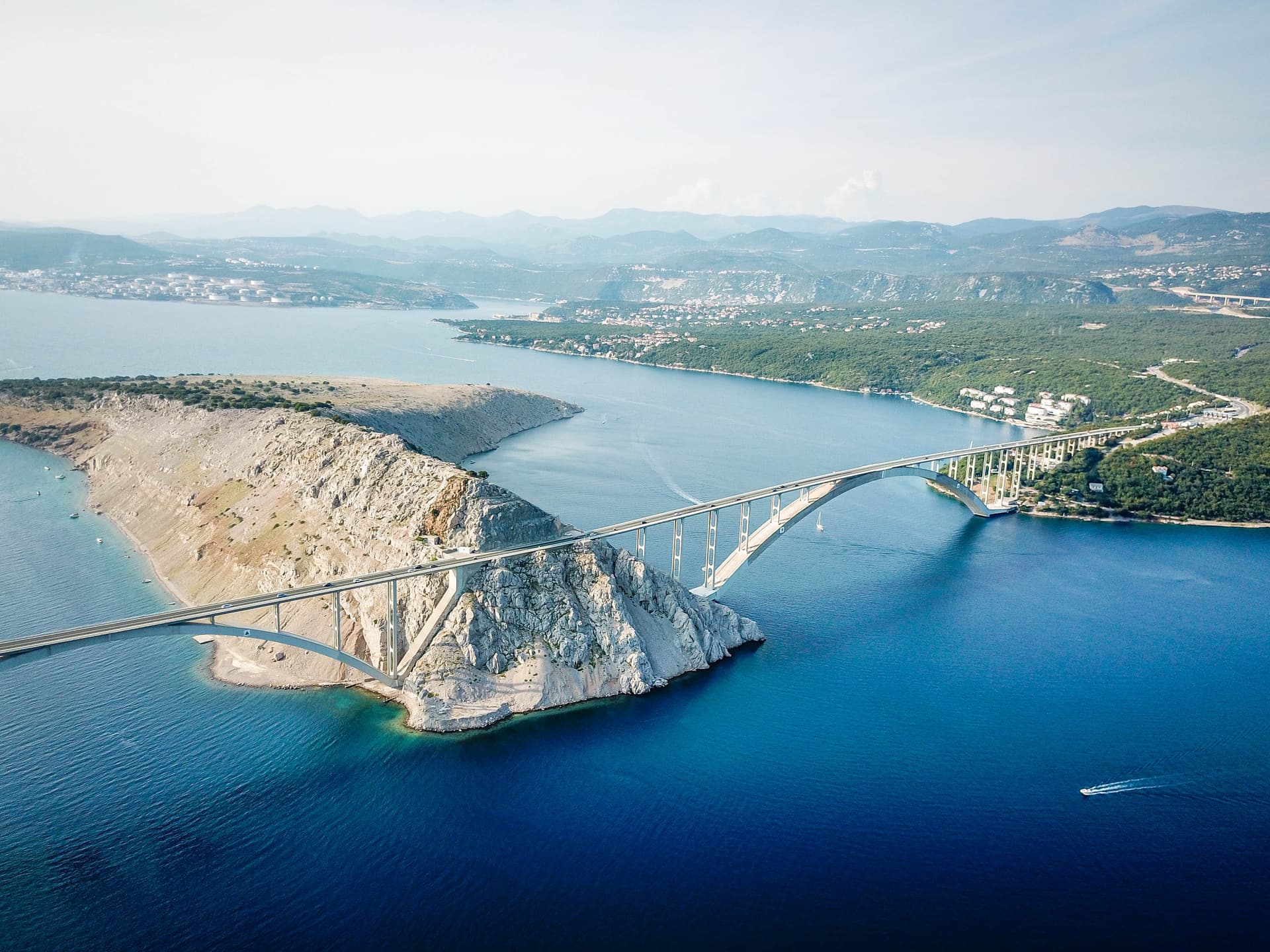 Arch bridge spanning deep blue sea connecting rocky island to green coastline with distant mountains.