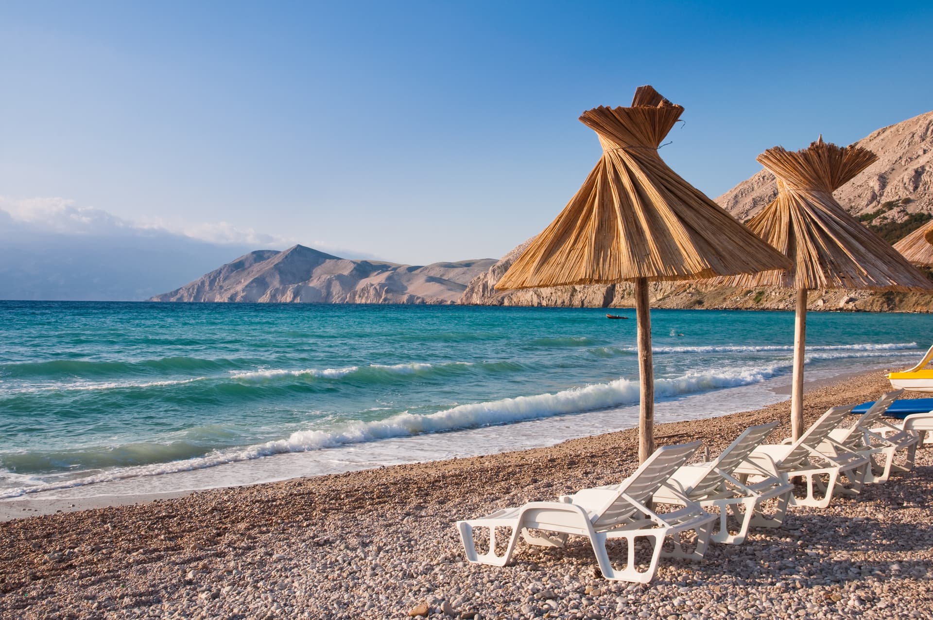 Straw beach umbrellas and white loungers on a pebble beach overlooking turquoise water and mountains. KRK.
