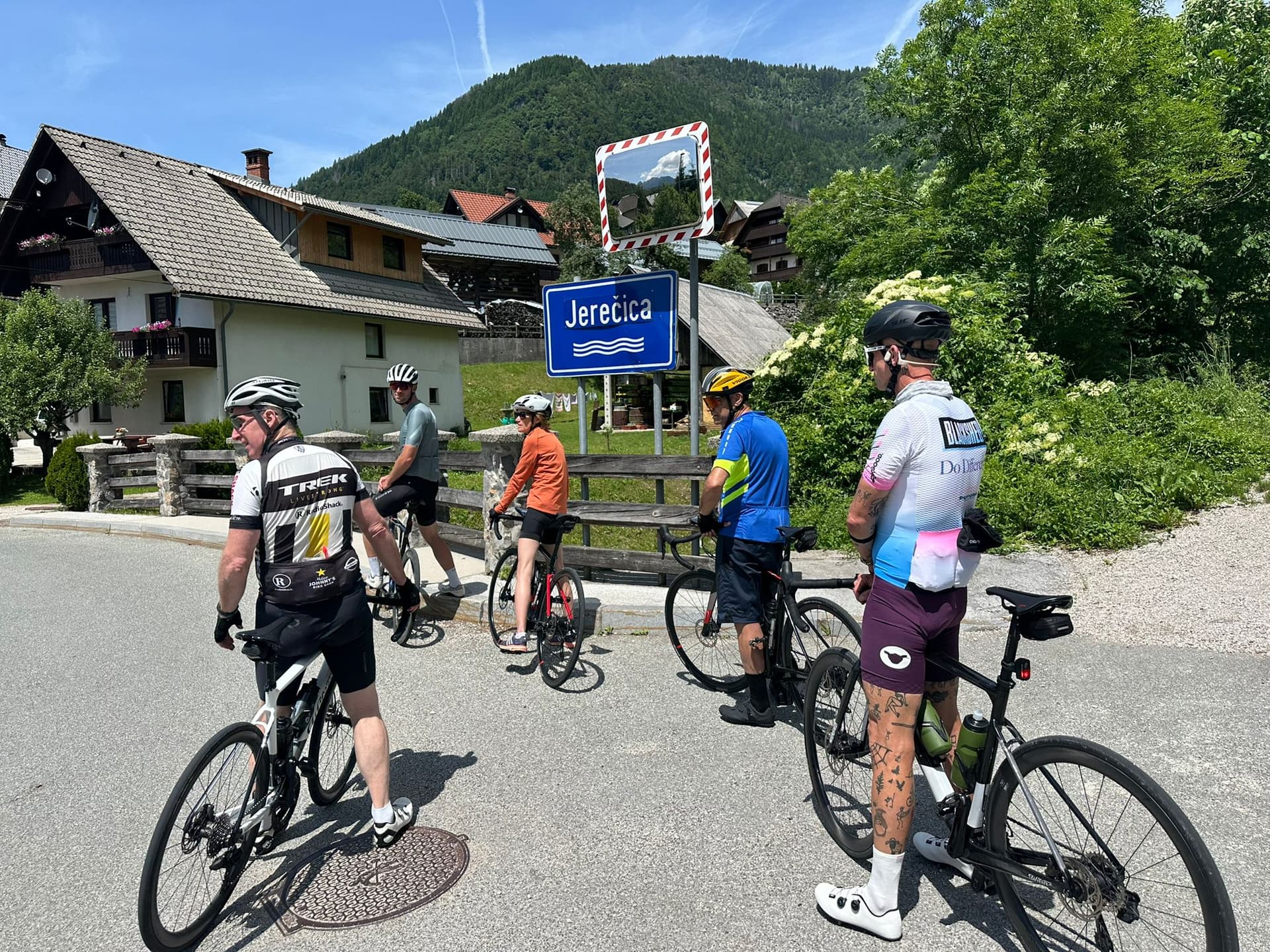 Cyclists stopped at Jerečica sign with houses and green mountains in the background.