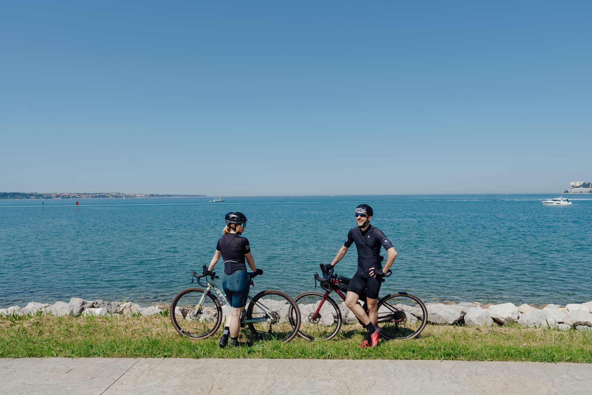 Cyclists resting with gravel bikes by the sea with a town visible across the water.
