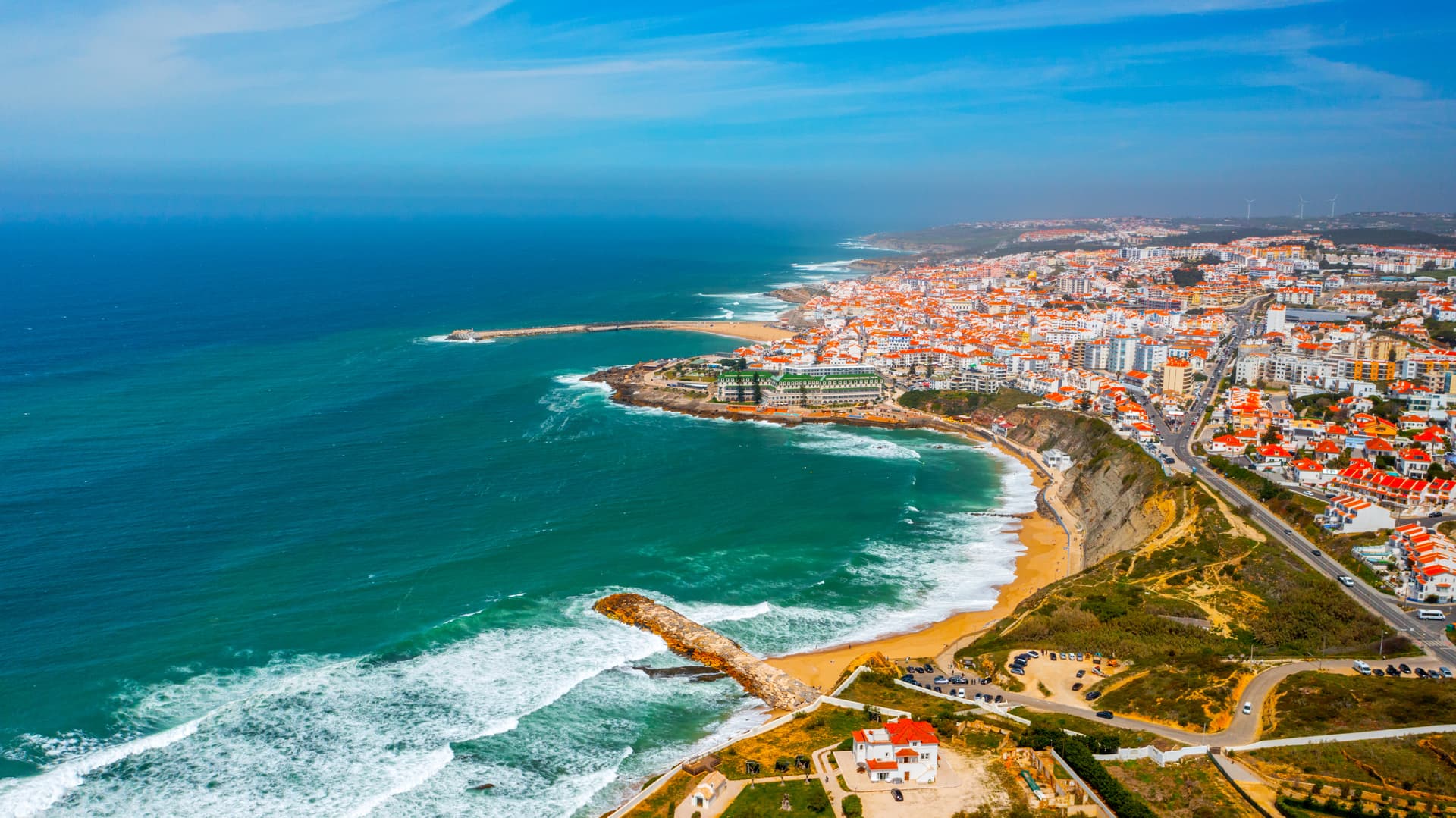 Aerial view of Ericeira coastal town with white buildings, orange roofs, and turquoise ocean waves.