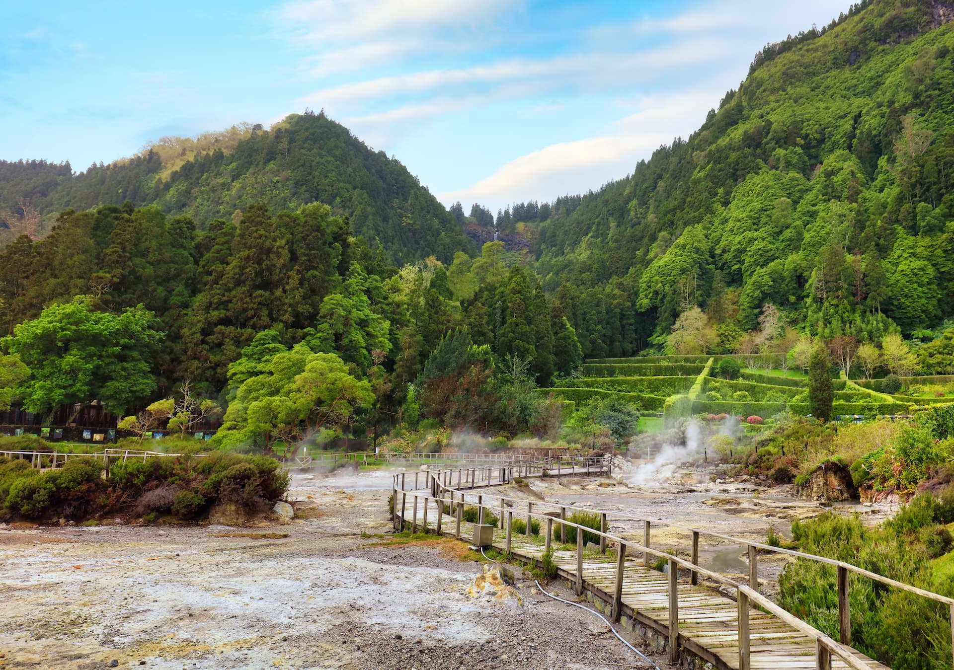 Wooden boardwalk winds through steaming geothermal area near lush green mountains of Furnas.