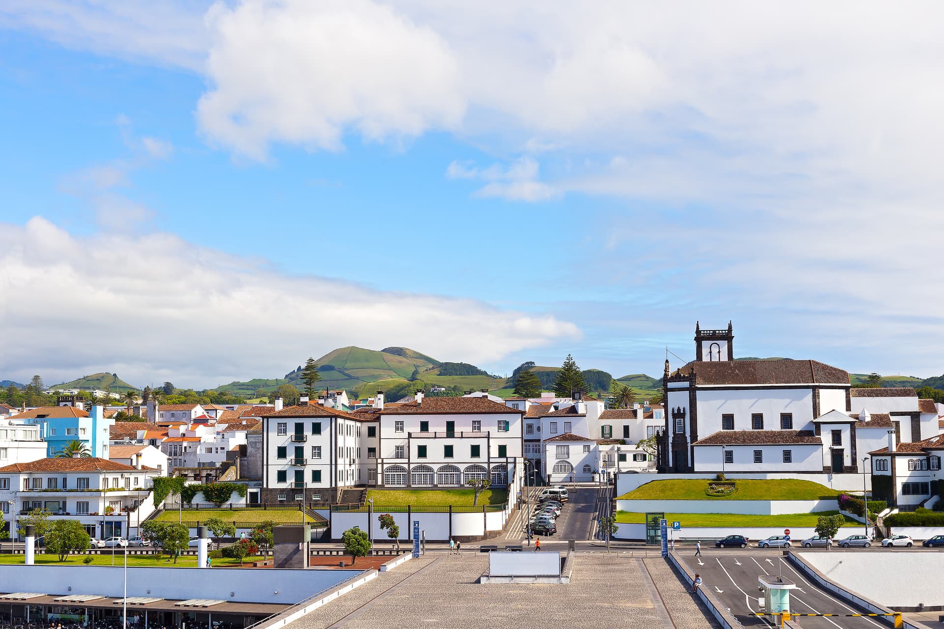 White buildings and church in Ponta Delgada with green hills under a blue sky.