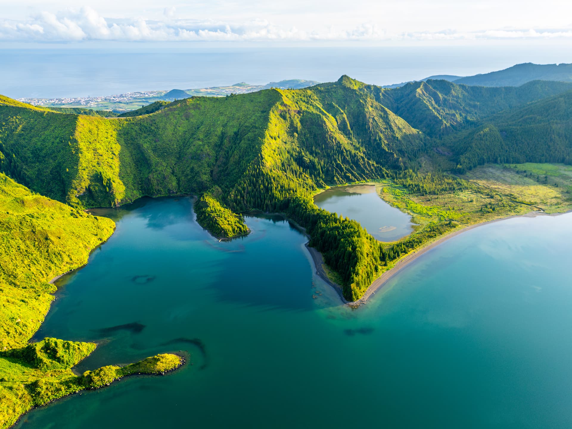 Aerial view of Lagoa do Fogo crater lake with lush green slopes and distant ocean view.