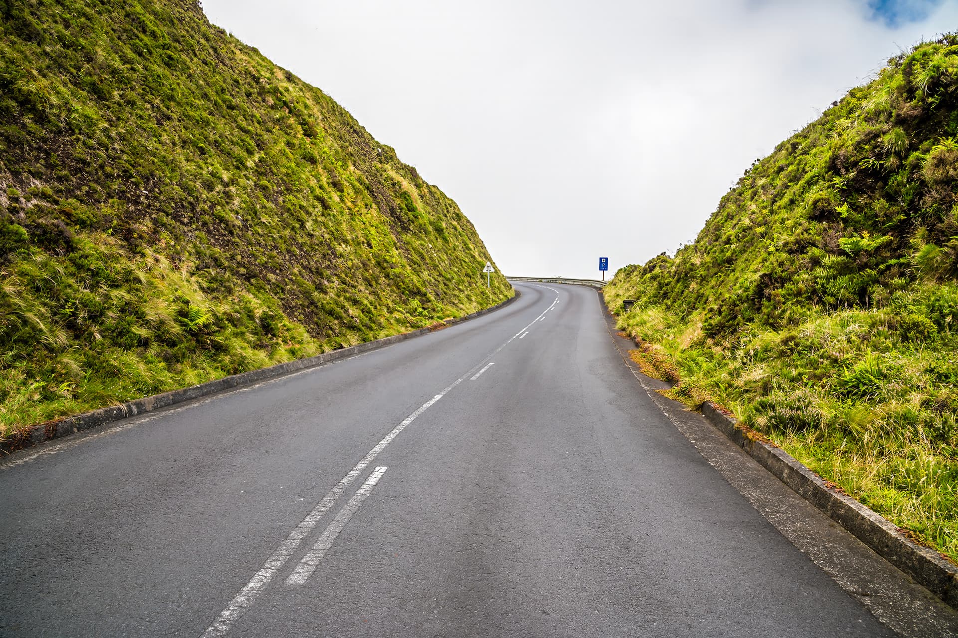 Winding asphalt road ascending between steep, lush green hillsides toward Lagoa do Fogo.