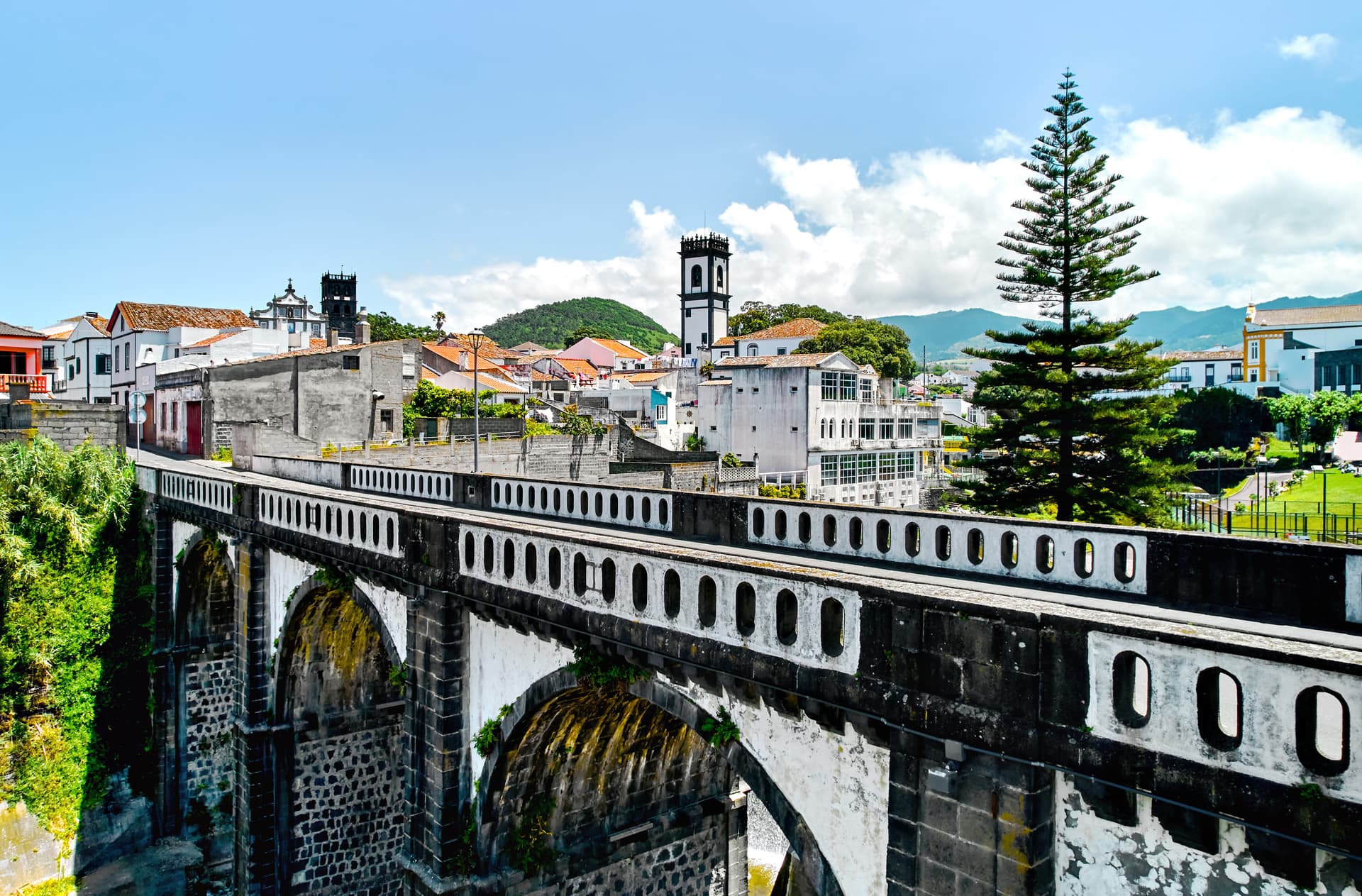 Stone bridge leading to Ribeira Grande town with white buildings and green mountains under blue sky.