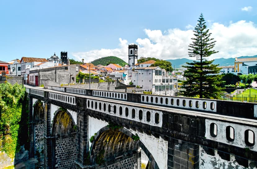 Stone bridge leading to Ribeira Grande town with white buildings and green mountains under blue sky.