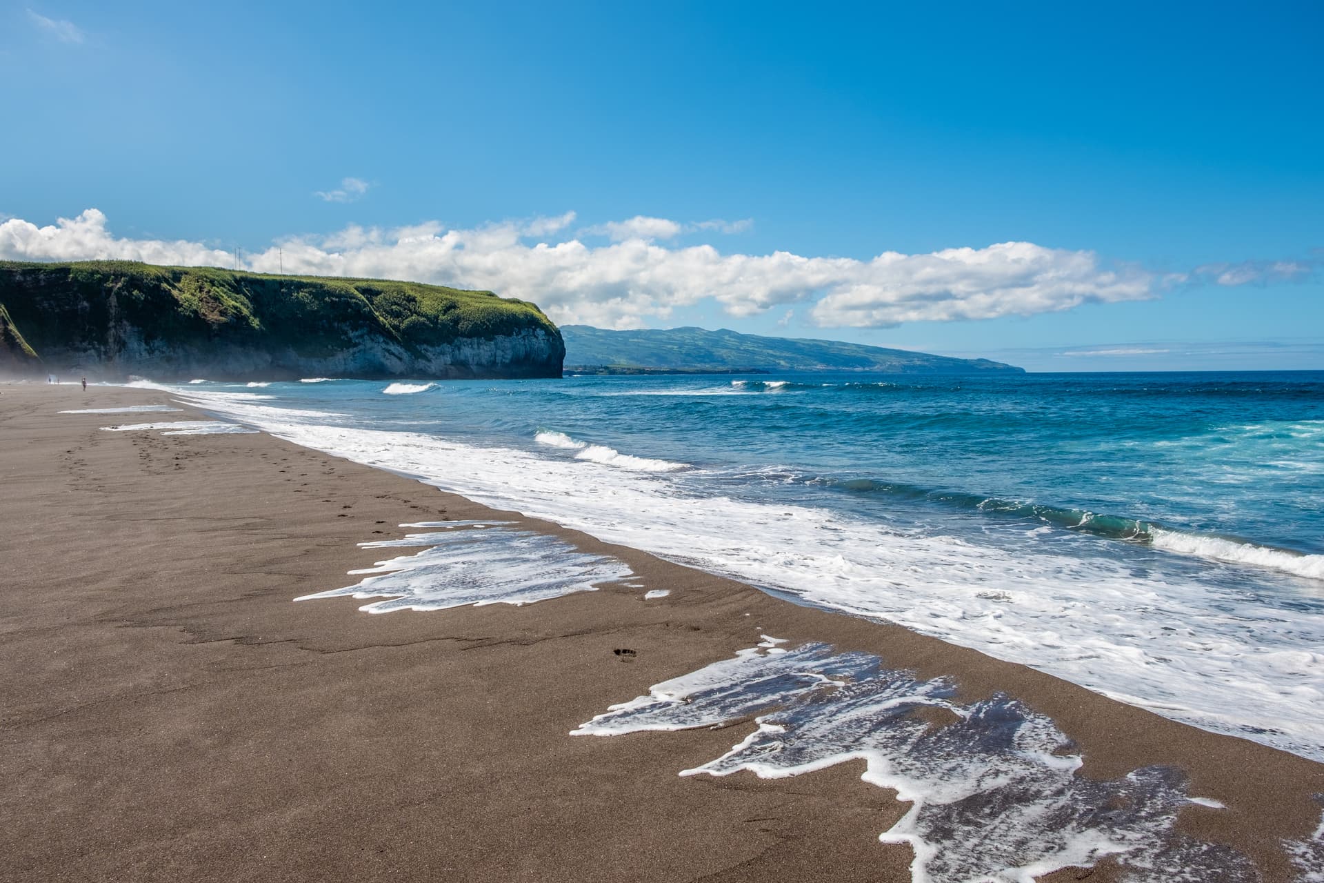 Dark sand beach at Santa Barbara, Ribeira Grande, with waves washing ashore under a blue sky.