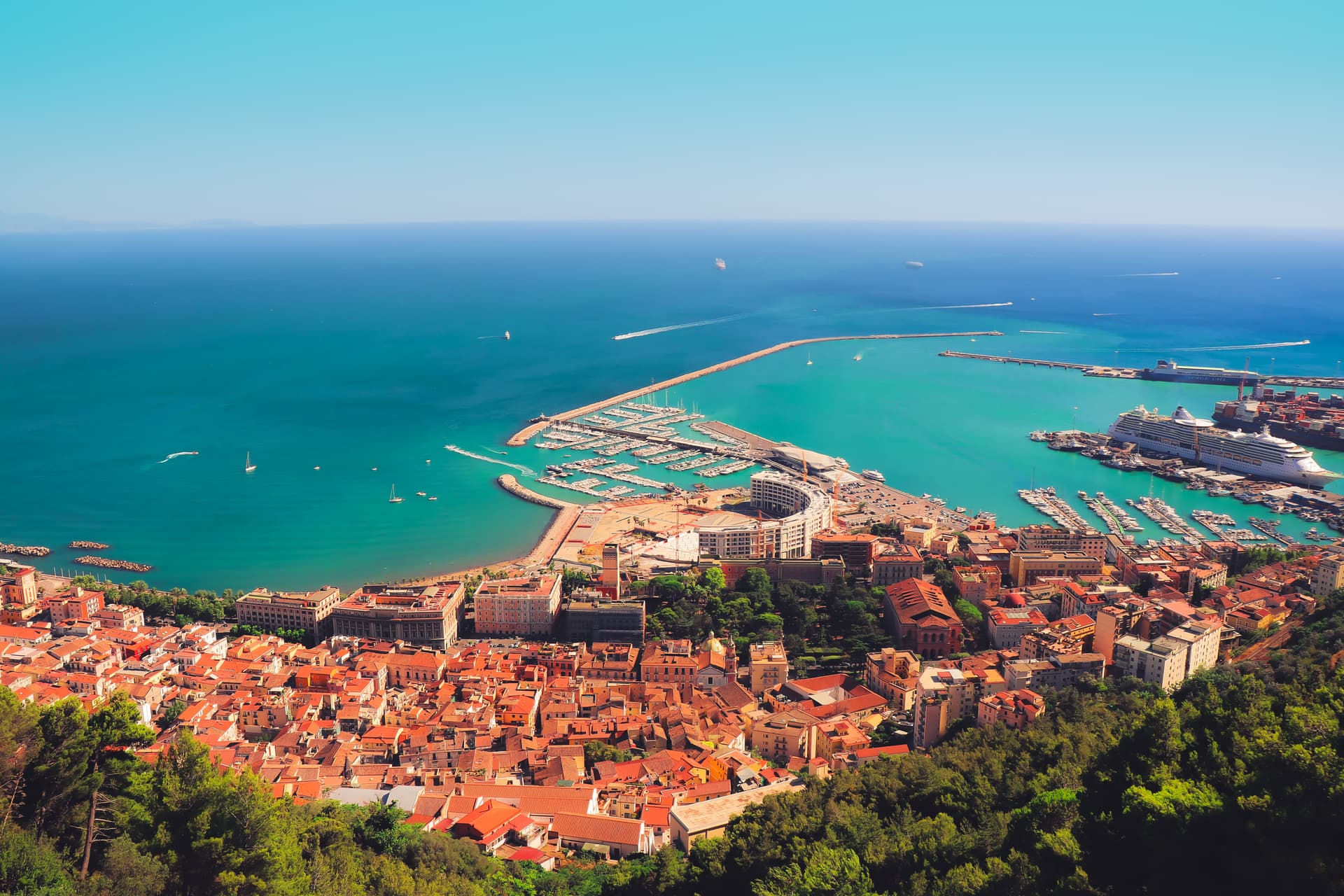 Aerial view of Salerno city with terracotta roofs overlooking a large harbor and turquoise Mediterranean Sea.