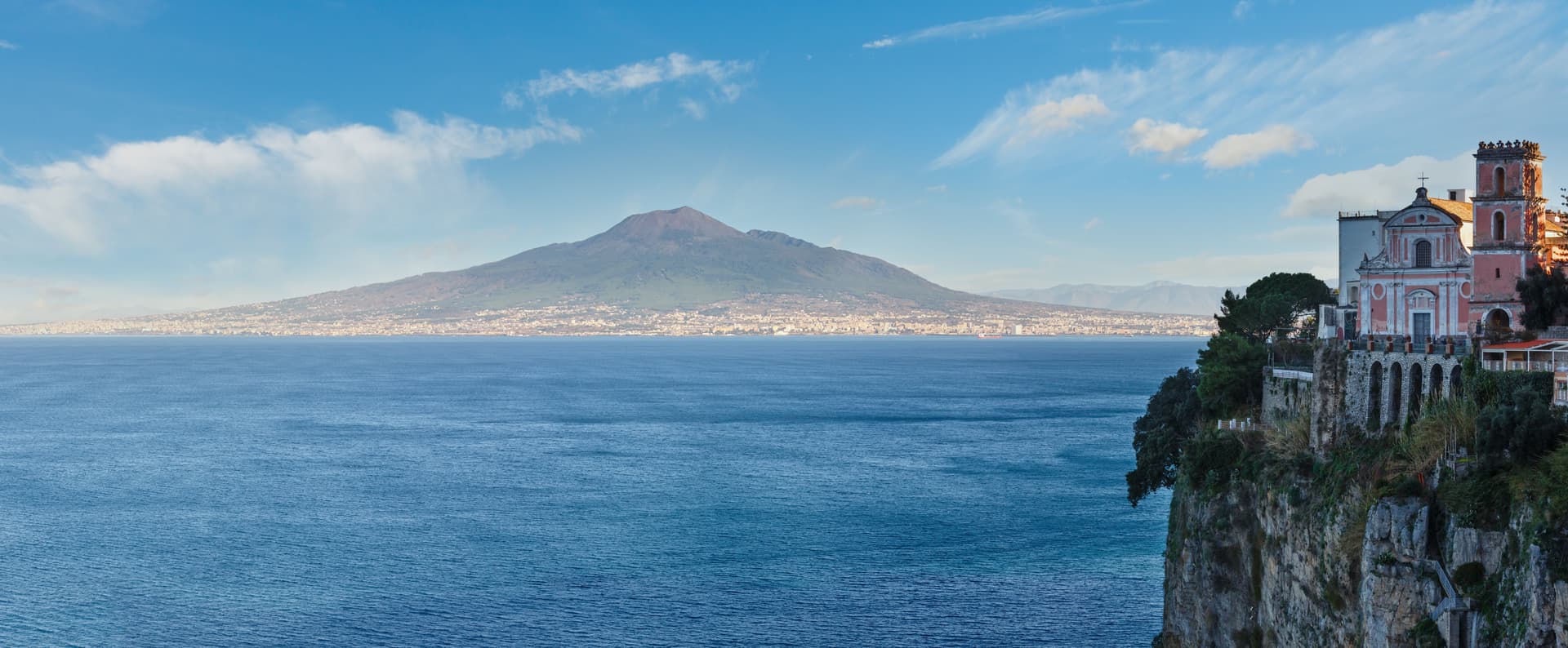 Mount Vesuvius looms over blue sea, with a church on a cliff edge.