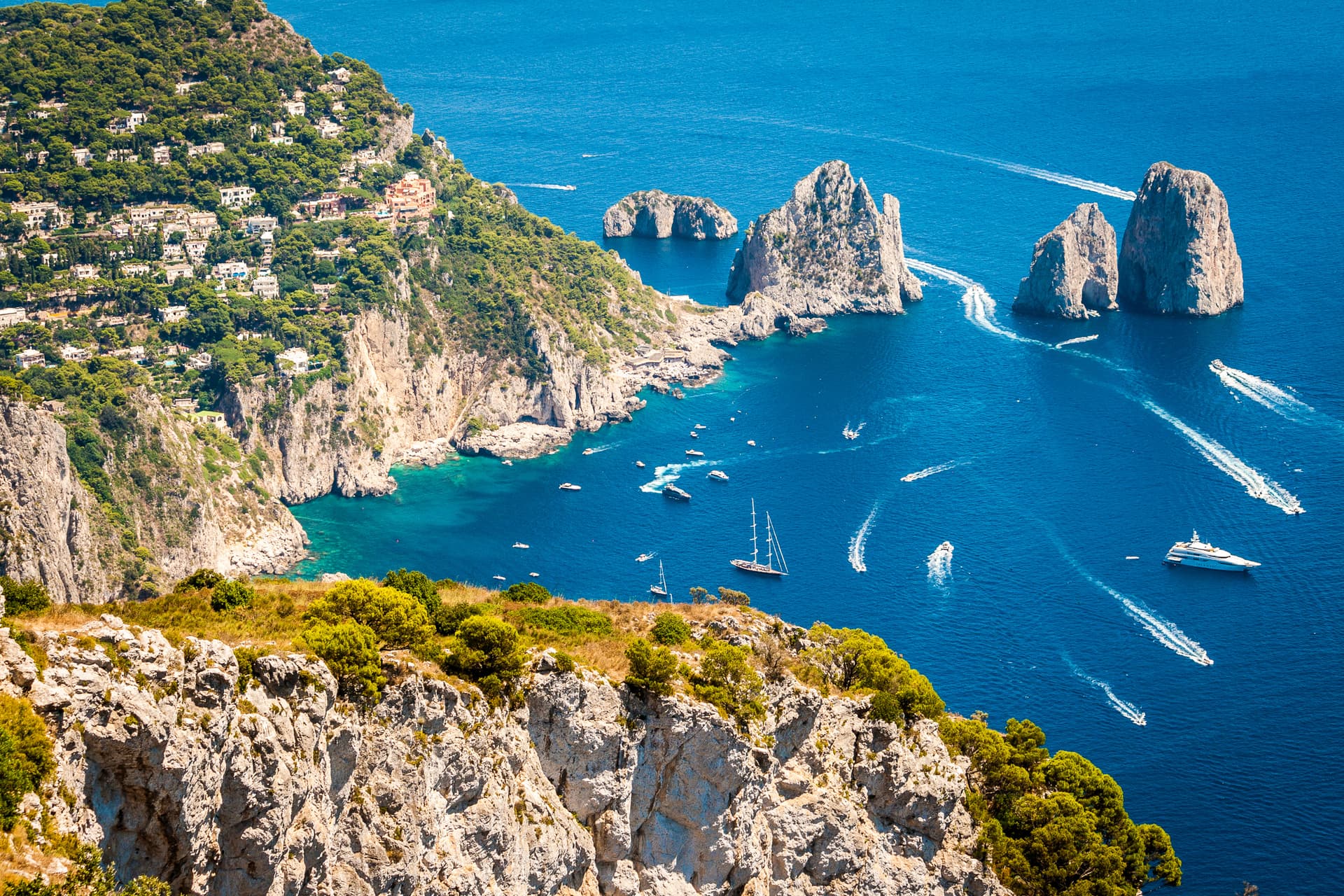 Birds-eye view of Island of Capri cliffs, Faraglioni rocks, and boats on deep blue sea.