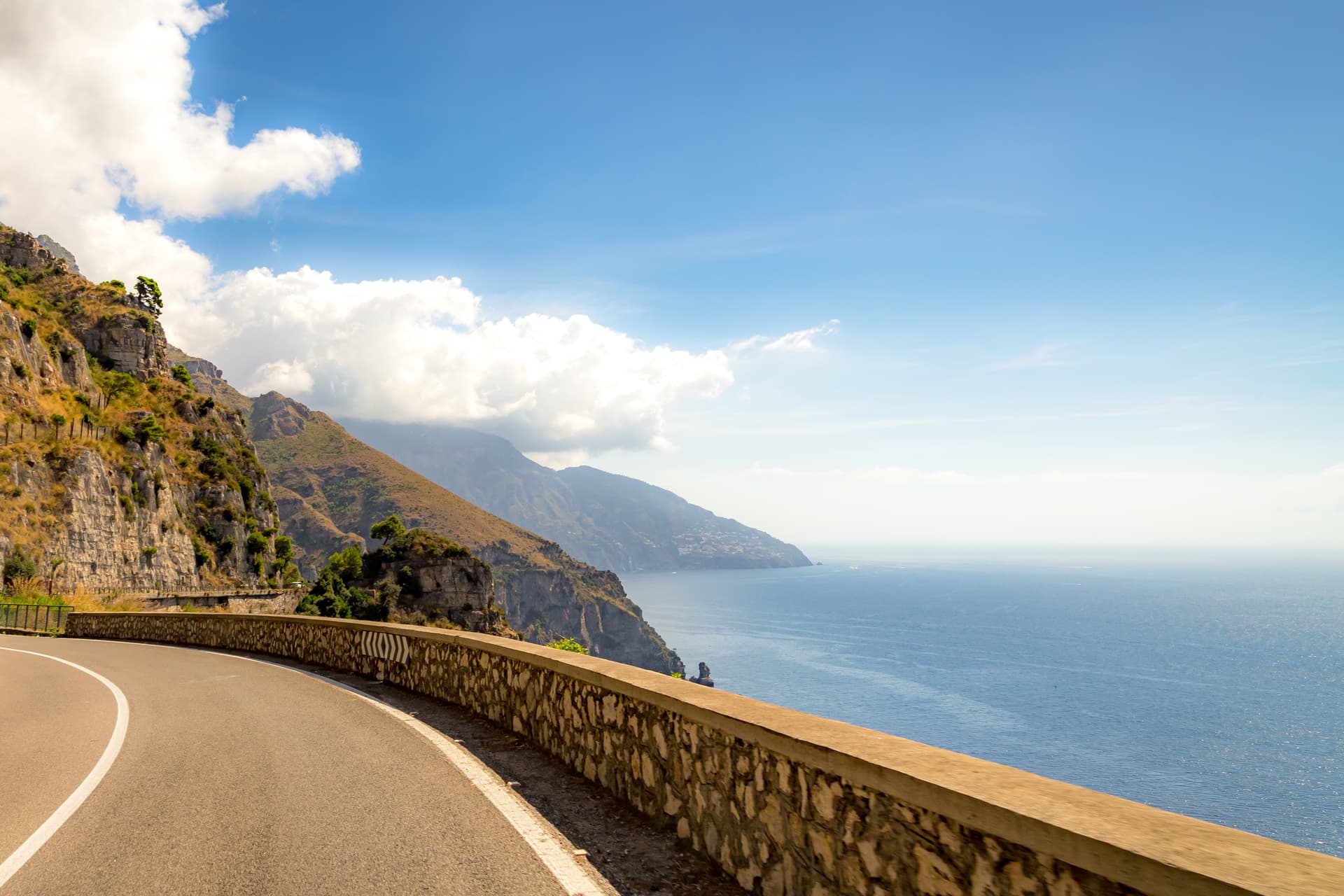 Winding coastal road along steep cliffs overlooking the bright blue Mediterranean Sea under a cloudy sky.