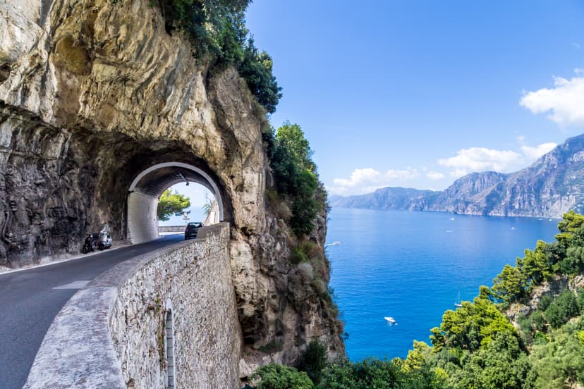 Coastal road tunnel carved into cliffside overlooking bright blue sea and mountains, Amalfi Coast.