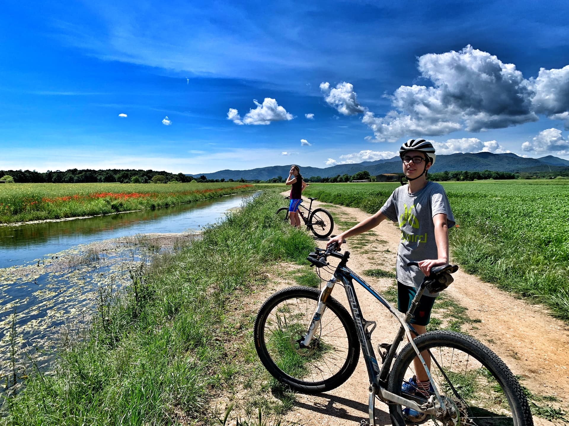 Cyclists on a dirt path beside a canal and green fields near Girona, Spain.
