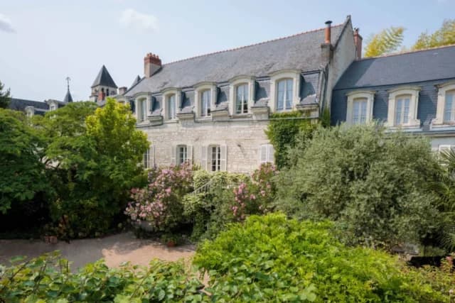 Stone building facade with a slate roof, surrounded by lush green trees and flowering bushes.