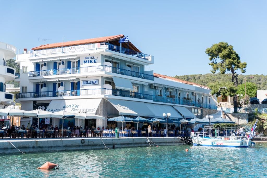 Hotel Mike and restaurant with outdoor seating by the harbor in Greece with a boat docked in the water.