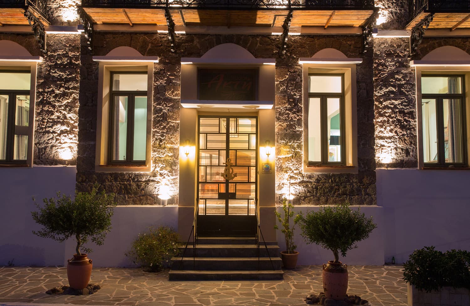 Illuminated stone building entrance with potted olive trees at night.