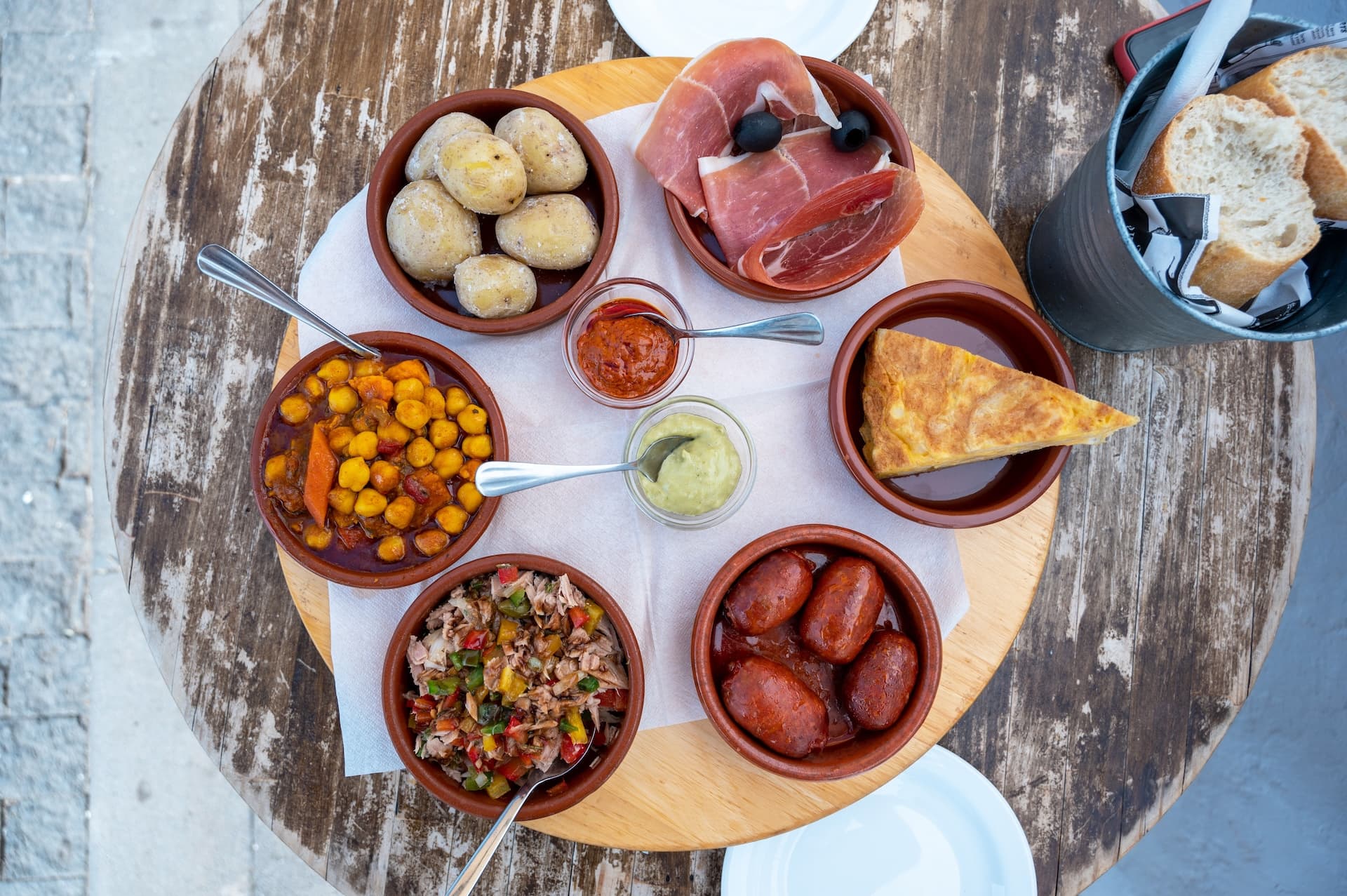Assortment of Spanish tapas including potatoes, chorizo, ham, and chickpea stew on a wooden serving board.