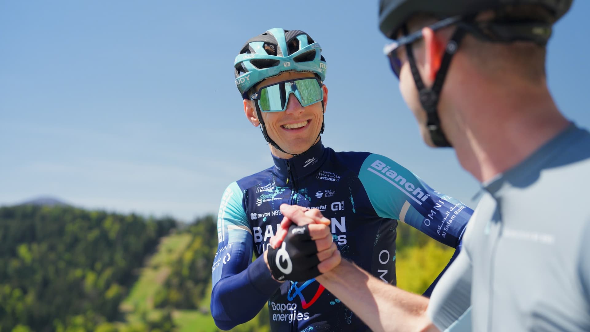 Two professional cyclists in team jerseys shaking hands outdoors with a blurred mountain forest background.