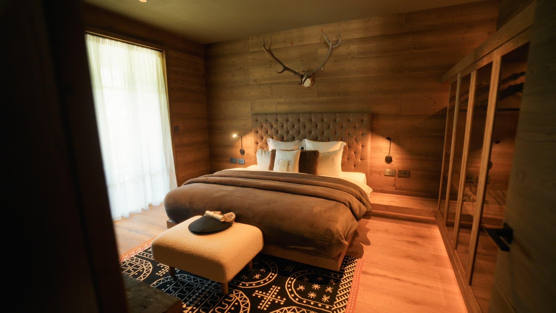 Chalet bedroom with wood-paneled walls, a tufted headboard, and deer antlers mounted above the bed.