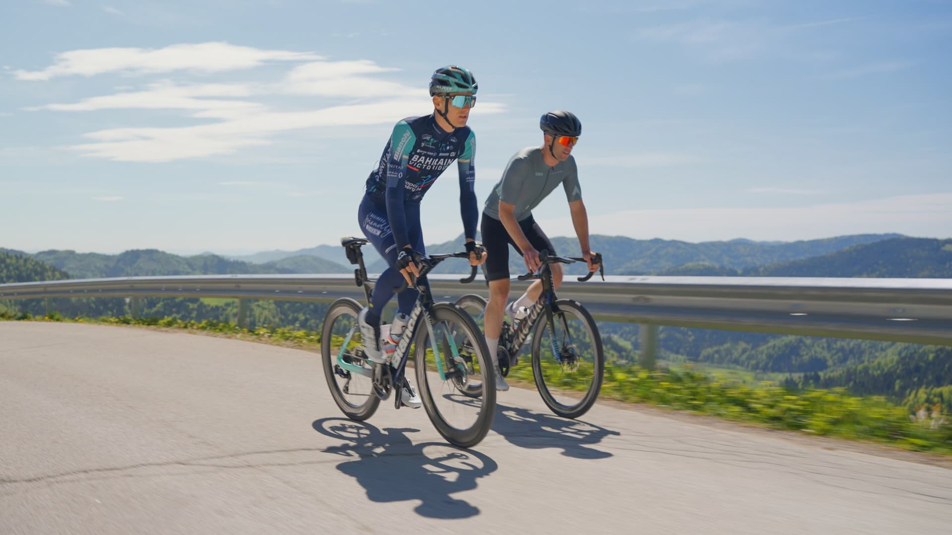 Two cyclists riding road bikes on a mountain pass with scenic hills in the background.
