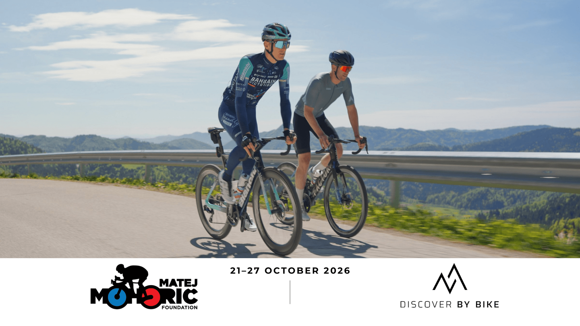 Two cyclists riding road bikes on a mountain road with forest and valley views.