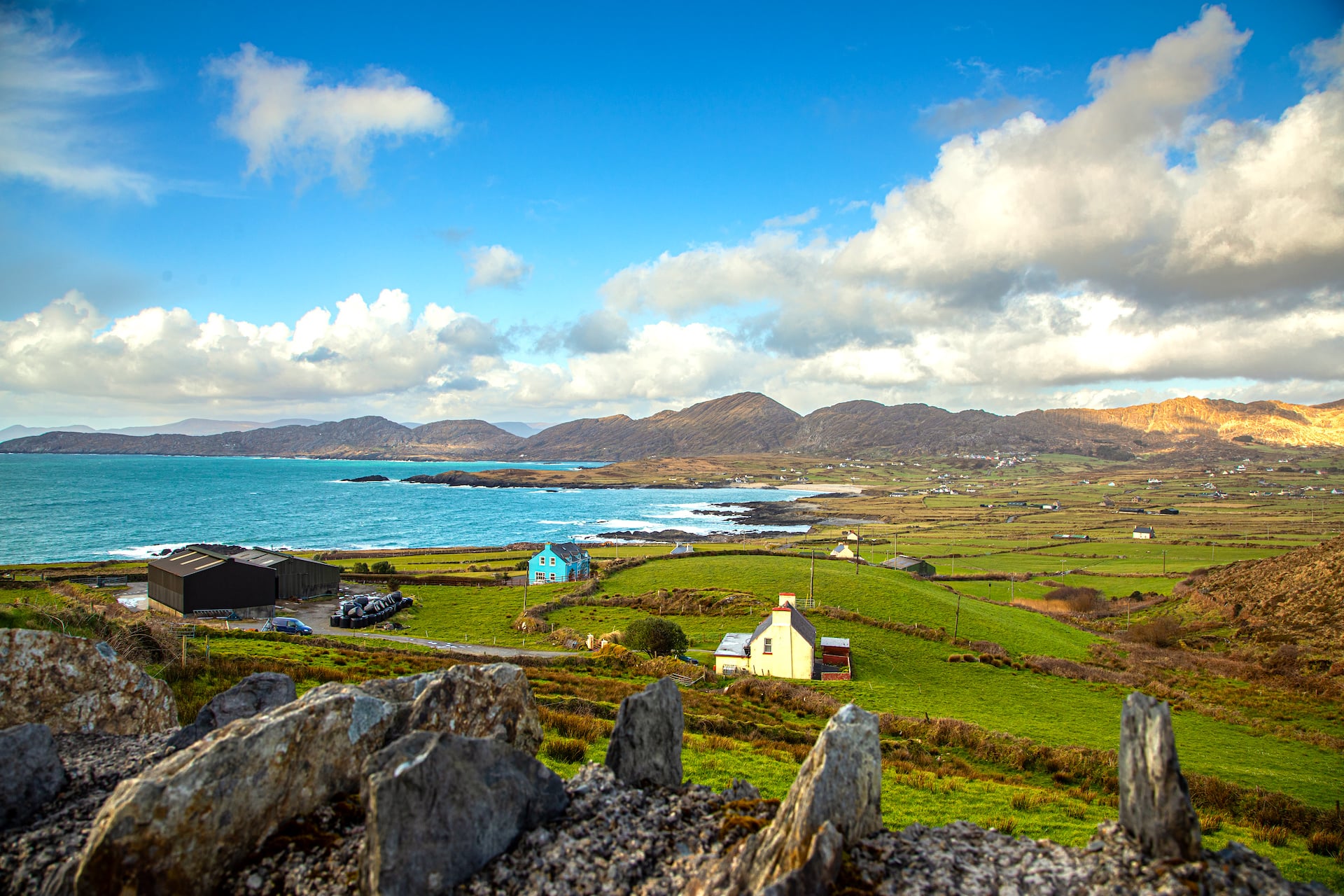 Coastal landscape with green fields, colorful houses, turquoise sea, and rolling hills under a cloudy blue sky.