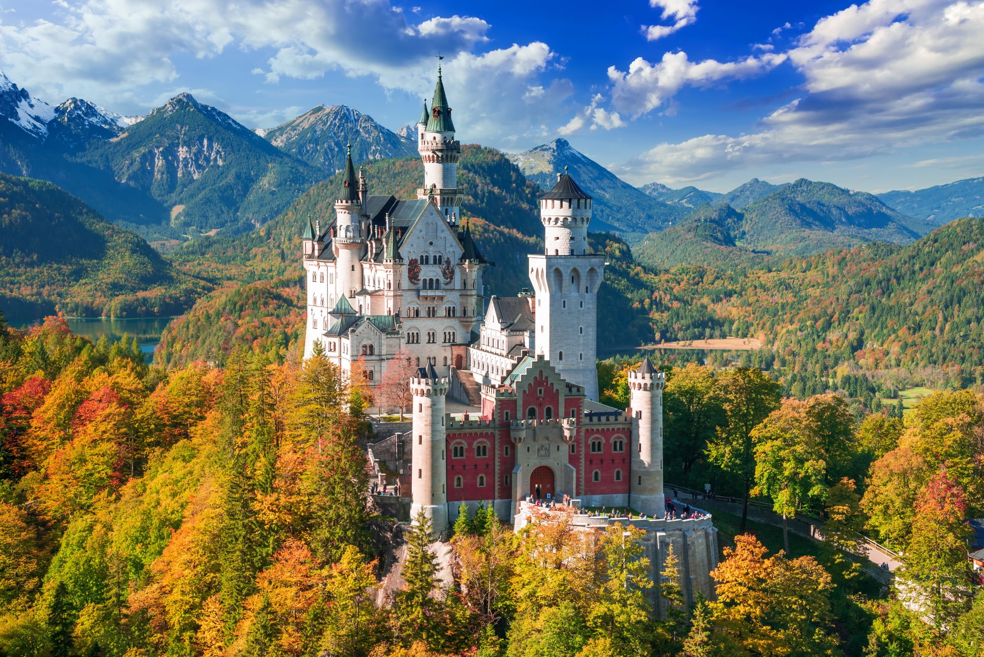 Neuschwanstein Castle with autumn foliage, mountains, and blue sky with clouds.