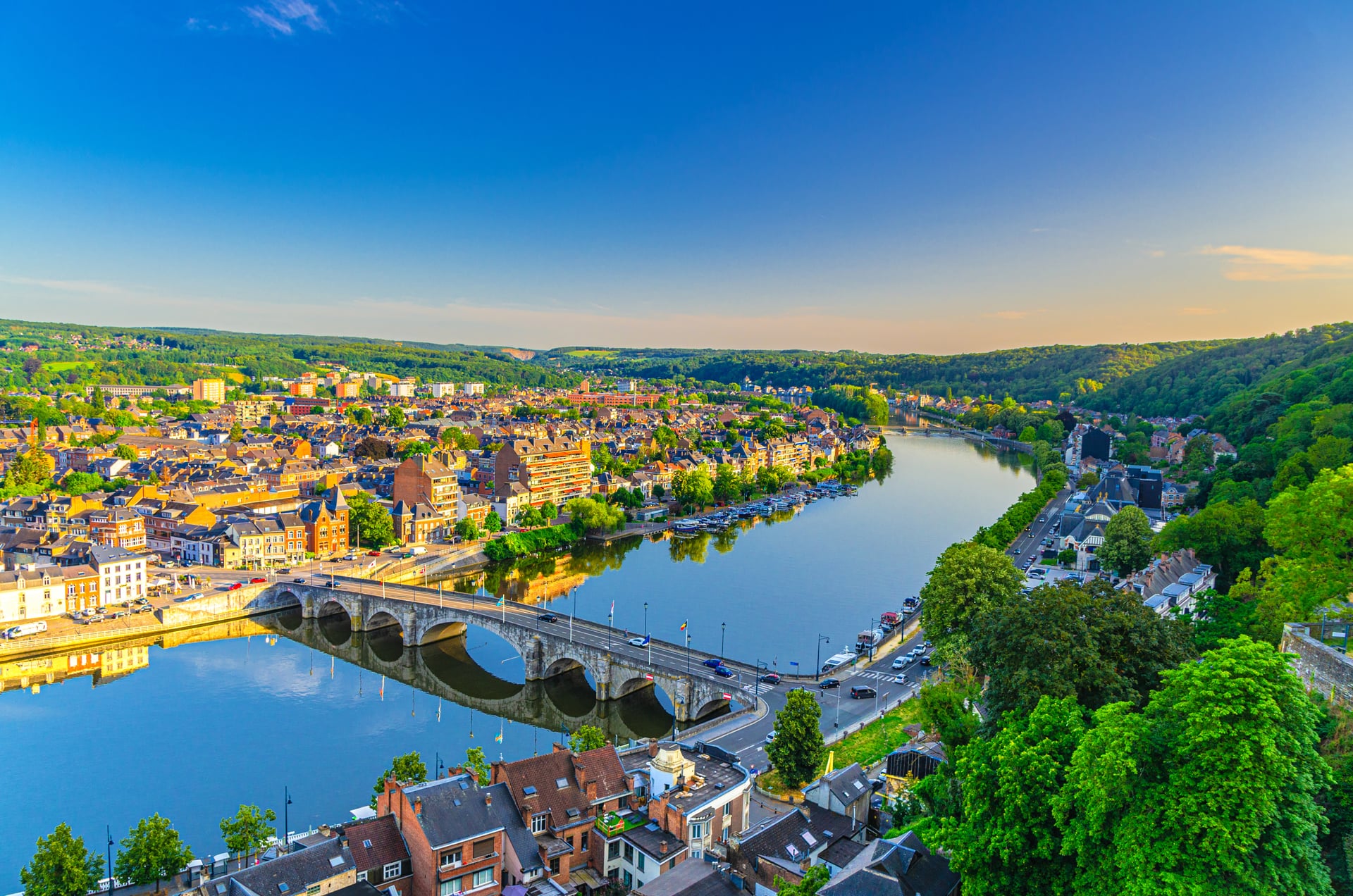 Cityscape with arched bridge over wide river reflecting buildings and lush green hills under blue sky.