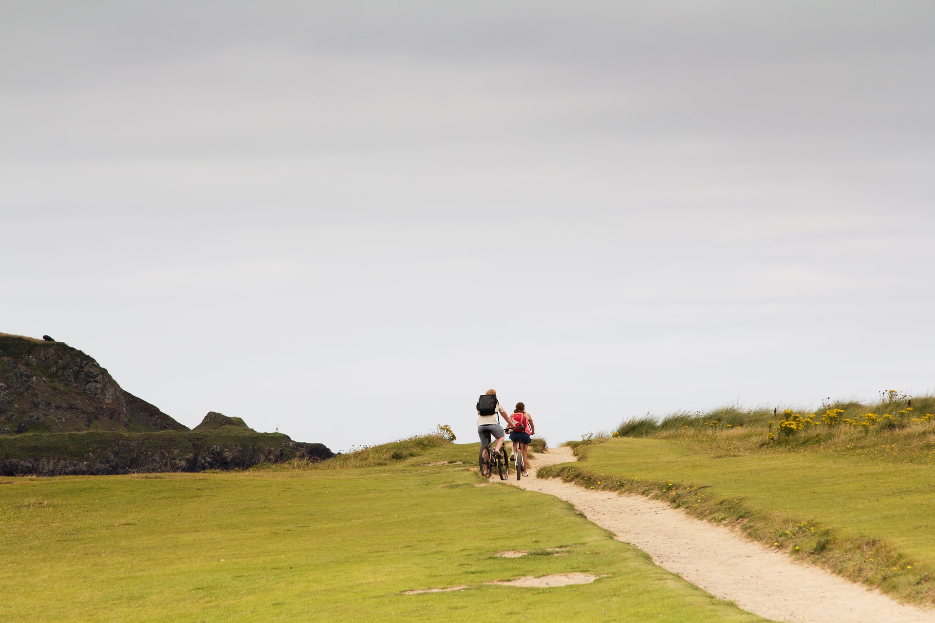 Two people cycling on a dirt path across grassy coastal hills under an overcast sky.