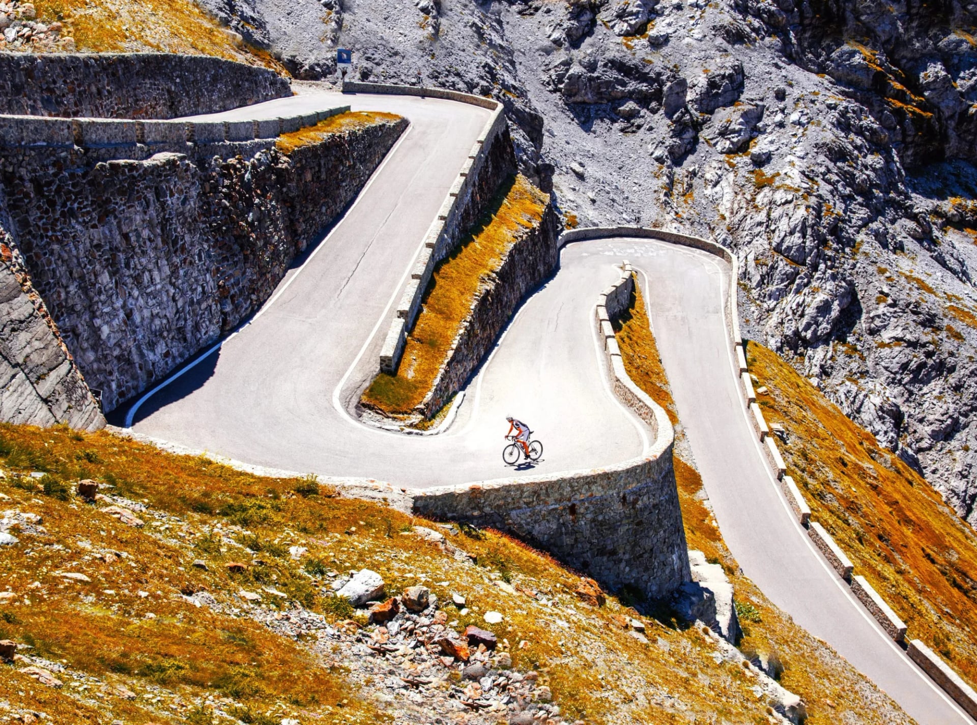 Cyclist ascending steep alpine switchback road with stone retaining walls and dry grass.