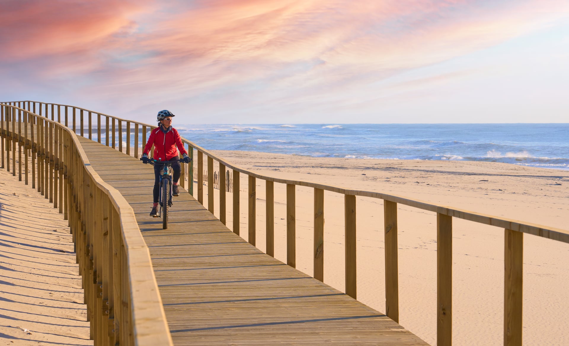 Cyclist on wooden boardwalk beside sandy beach and ocean under pink sunset sky in Portugal.