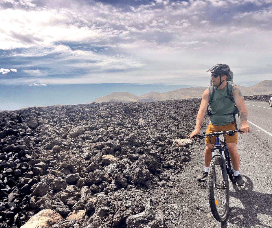Cyclist with backpack riding on a paved road beside a volcanic lava field with distant mountains.