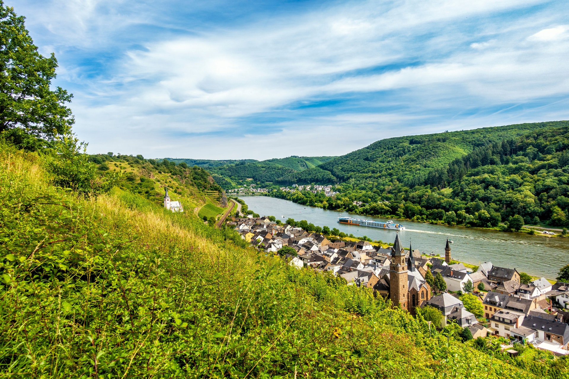River cruise ship passing village nestled between steep, green hillsides under blue sky.