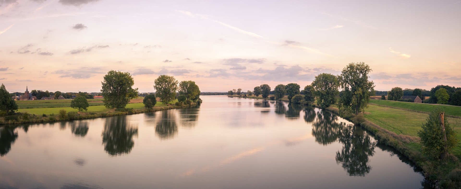 Calm river reflecting trees and soft sunset sky near a small village, Meuse River.