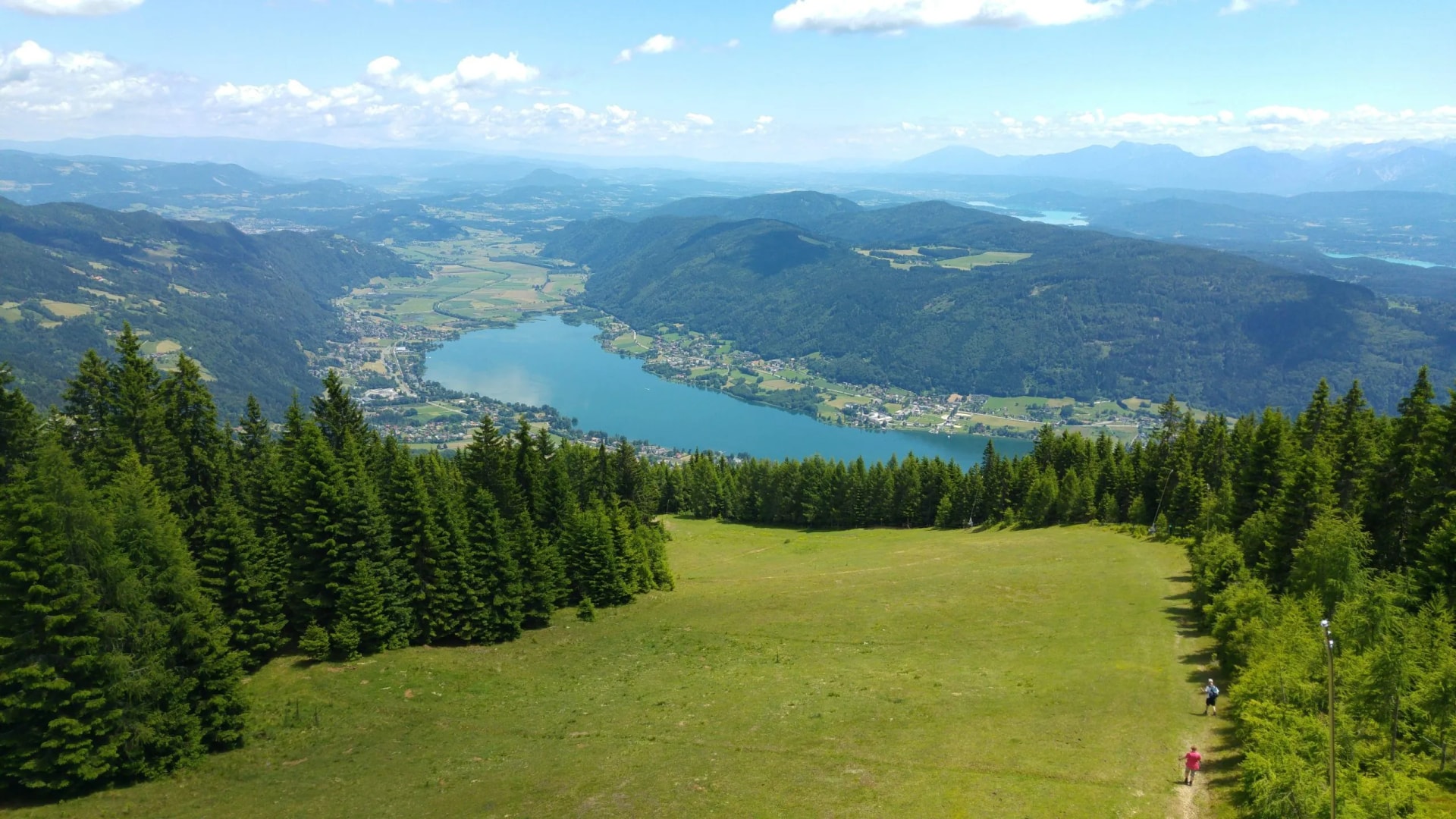 Hikers on grassy slope overlooking forested mountains and blue lake valley on sunny day