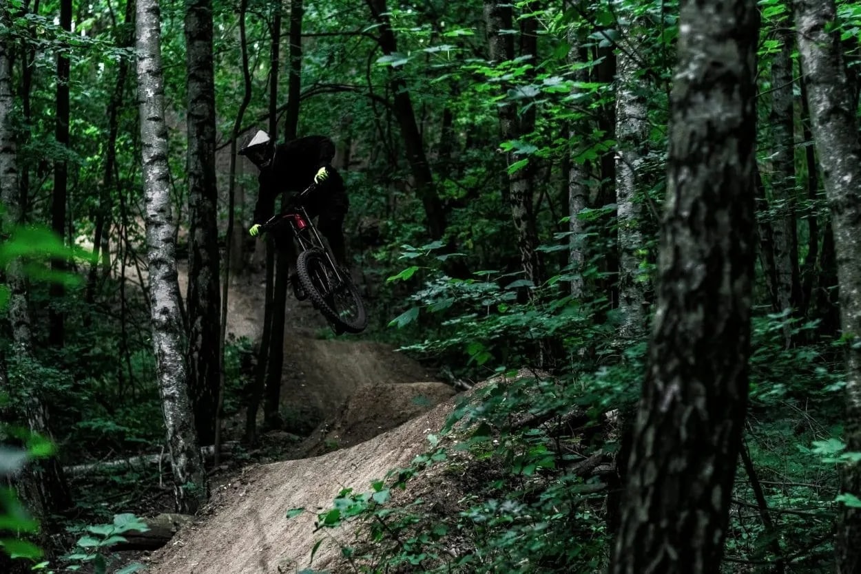 Mountain biker catching air off a dirt jump in a dense, dark forest setting