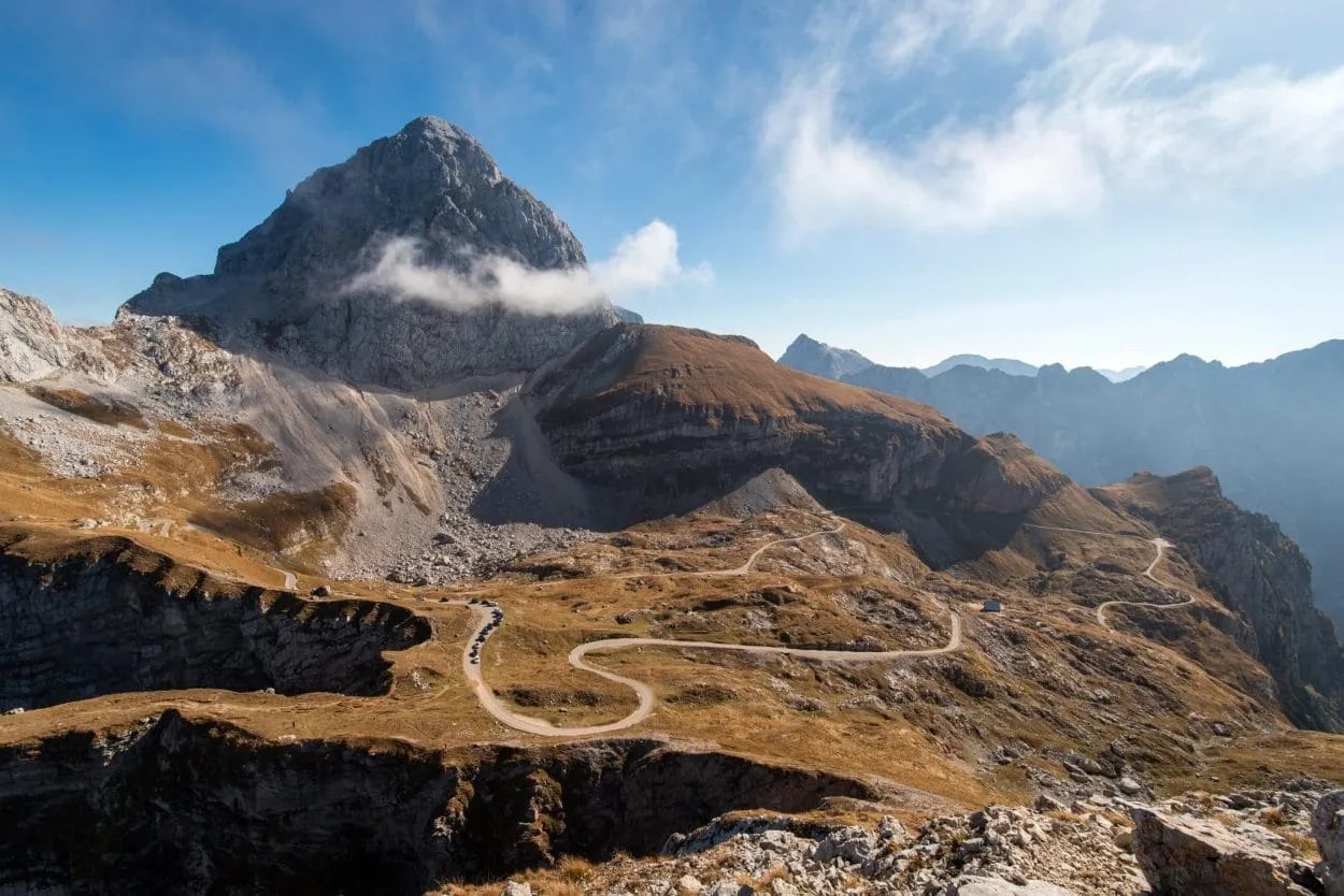 Winding mountain road traversing dry, grassy slopes below a rocky peak under a blue sky.