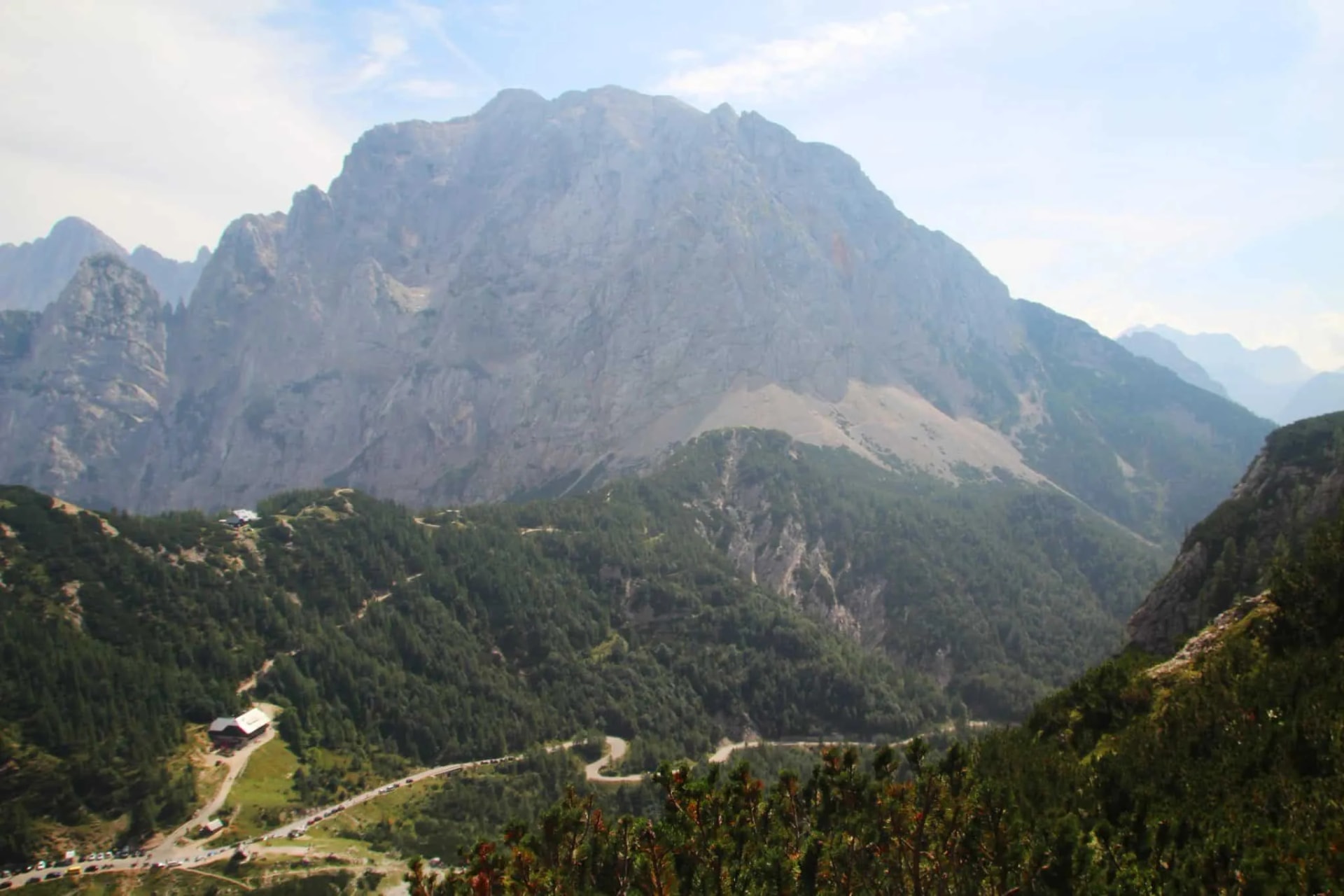 Winding mountain road below large gray peak with forested slopes, likely Vršič Pass.