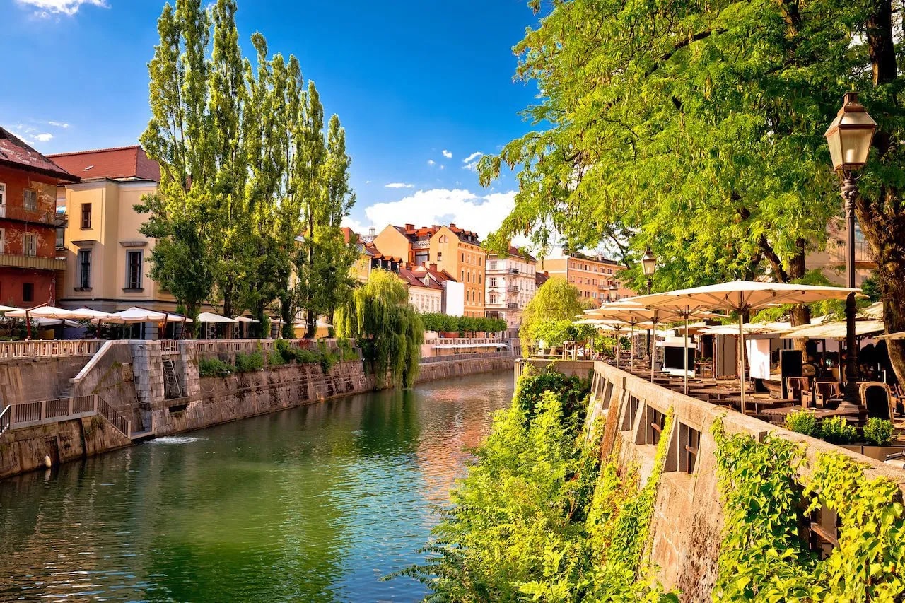 Riverfront cafe seating under umbrellas next to a canal in Ljubljana.